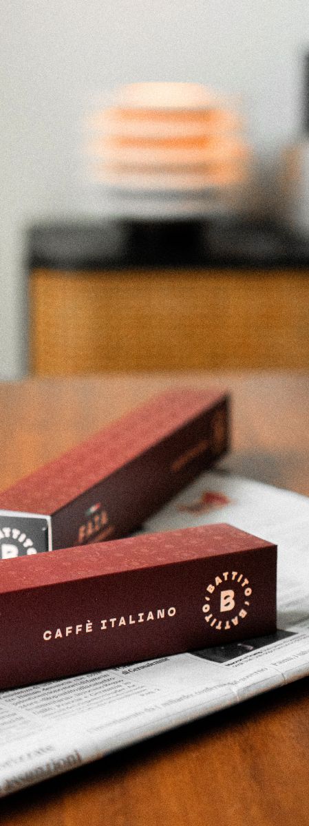 Twee doosjes Battito Faia koffiecapsules voor Nespresso-koffiecupmachines op een houten tafel met een krant en lamp op de achtergrond